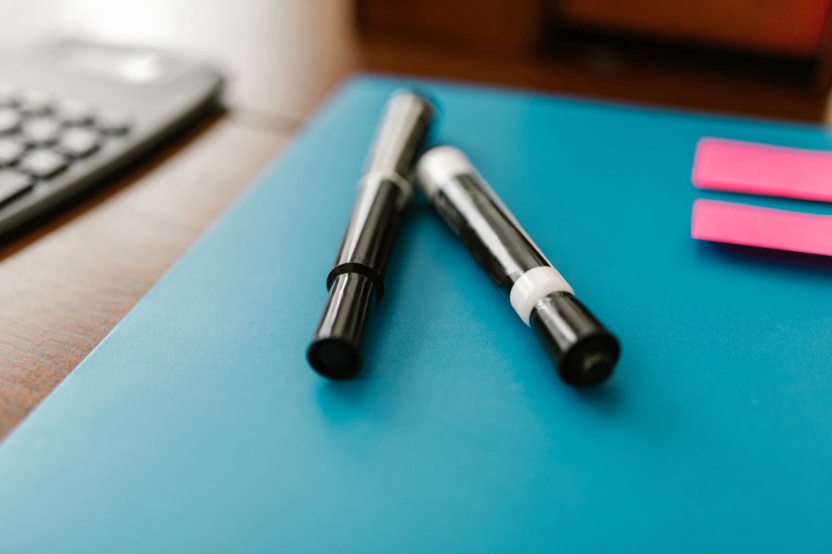 Two black pens lying on a closed blue folder on a desk, with a calculator and pink sticky notes at the edge