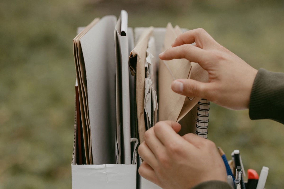 Hands pulling an envelope from a file box of papers, with a spiral notebook and pens beside it