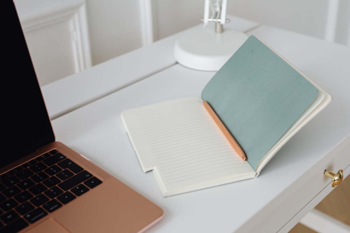 A rose-gold laptop with a dark screen beside an open notebook and a pencil on a white desk