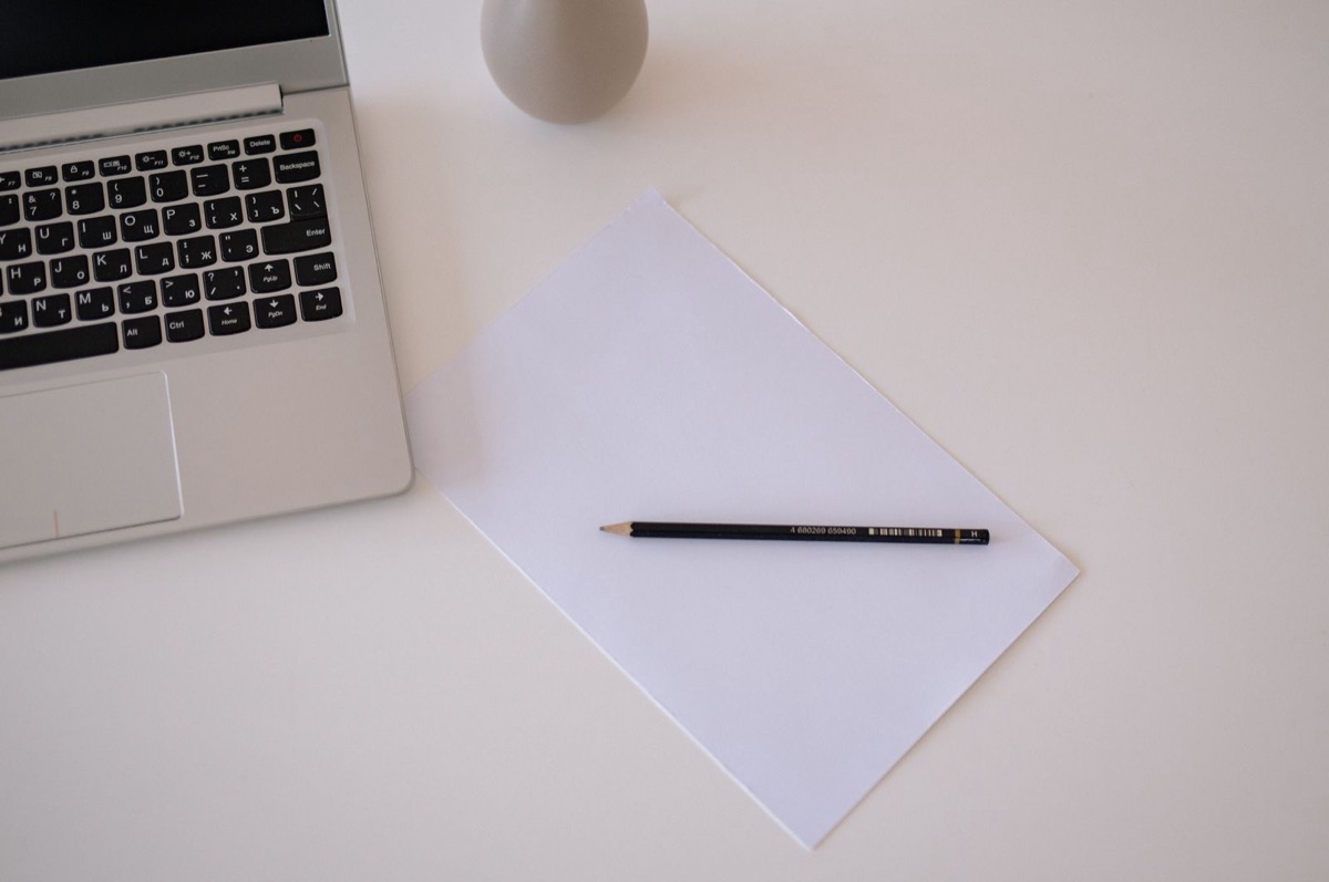 A blank sheet of paper with a pencil on a white desk, the corner of a laptop at the top left
