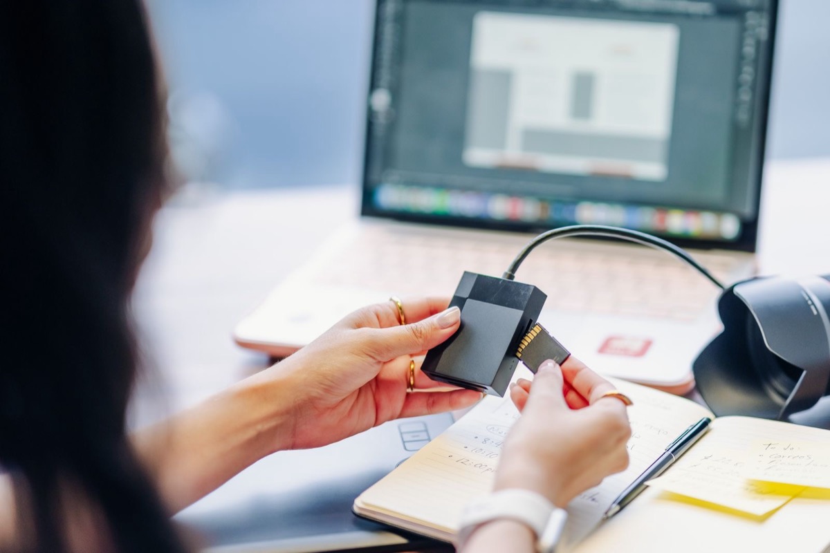 Hands holding a small black card reader beside an open notebook and a laptop