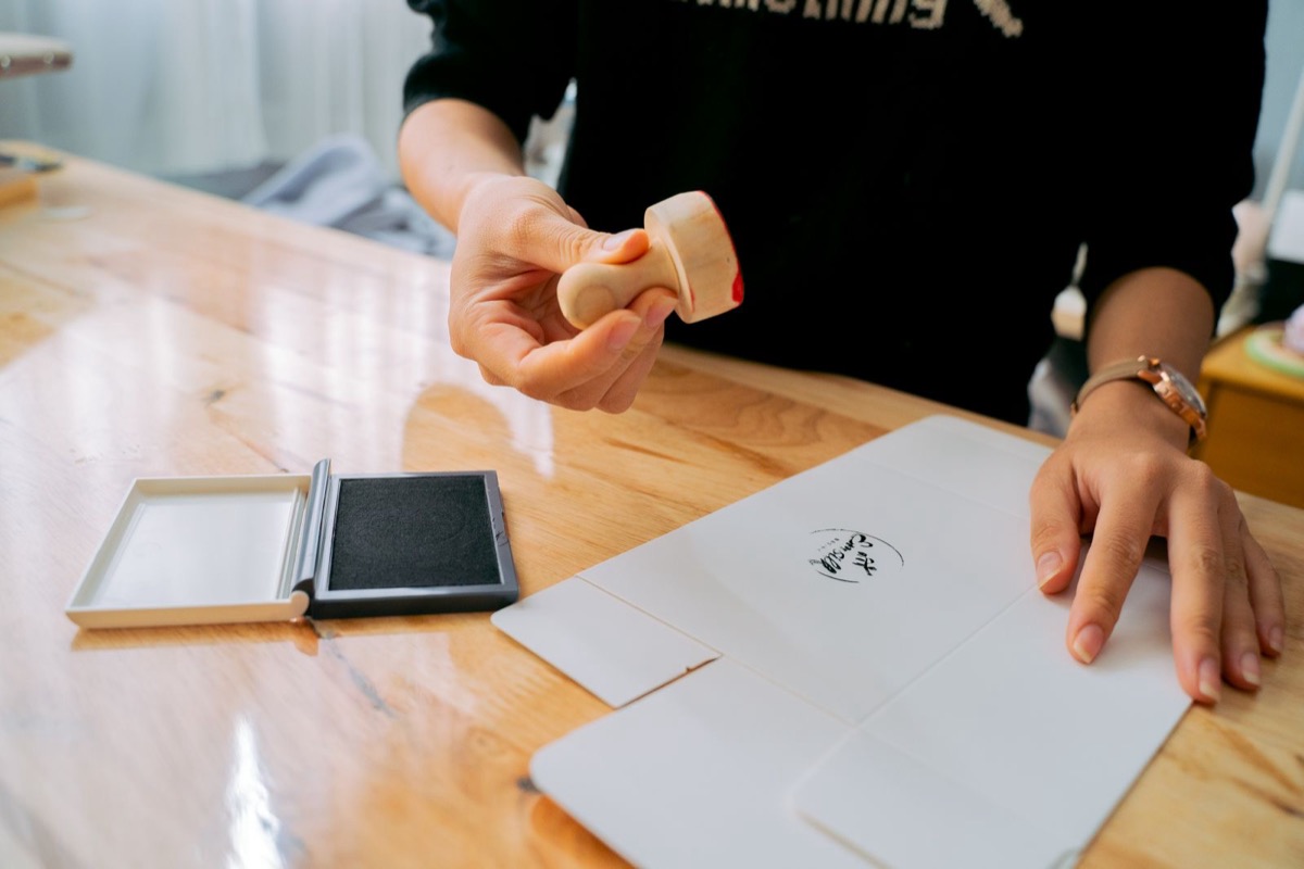 A hand holding a wooden stamp above a sheet with a small stamped mark, beside an open ink pad on a wooden counter