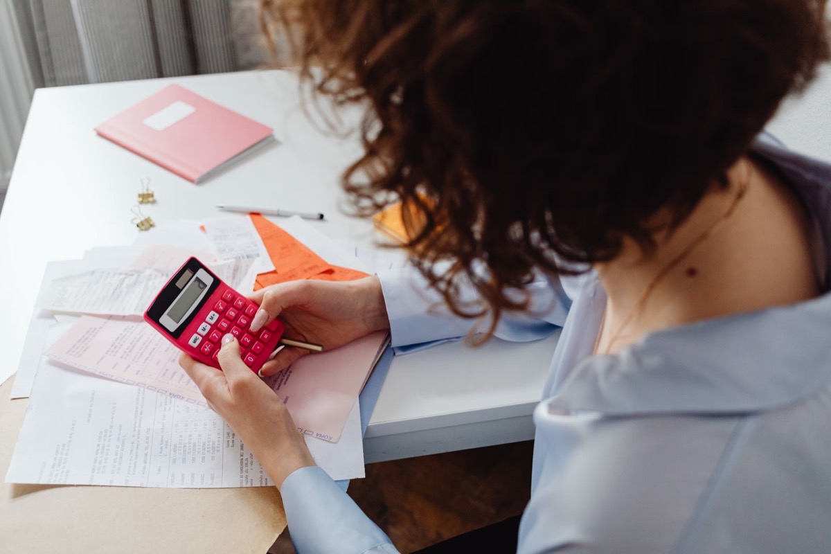 A woman seen from behind using a pink calculator over printed sheets and a pink notebook on a white desk