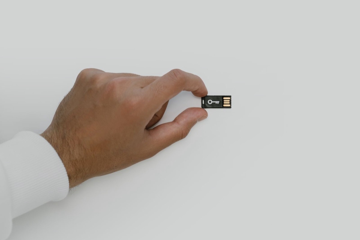 A hand holding a small USB security key with a key symbol against a plain light background