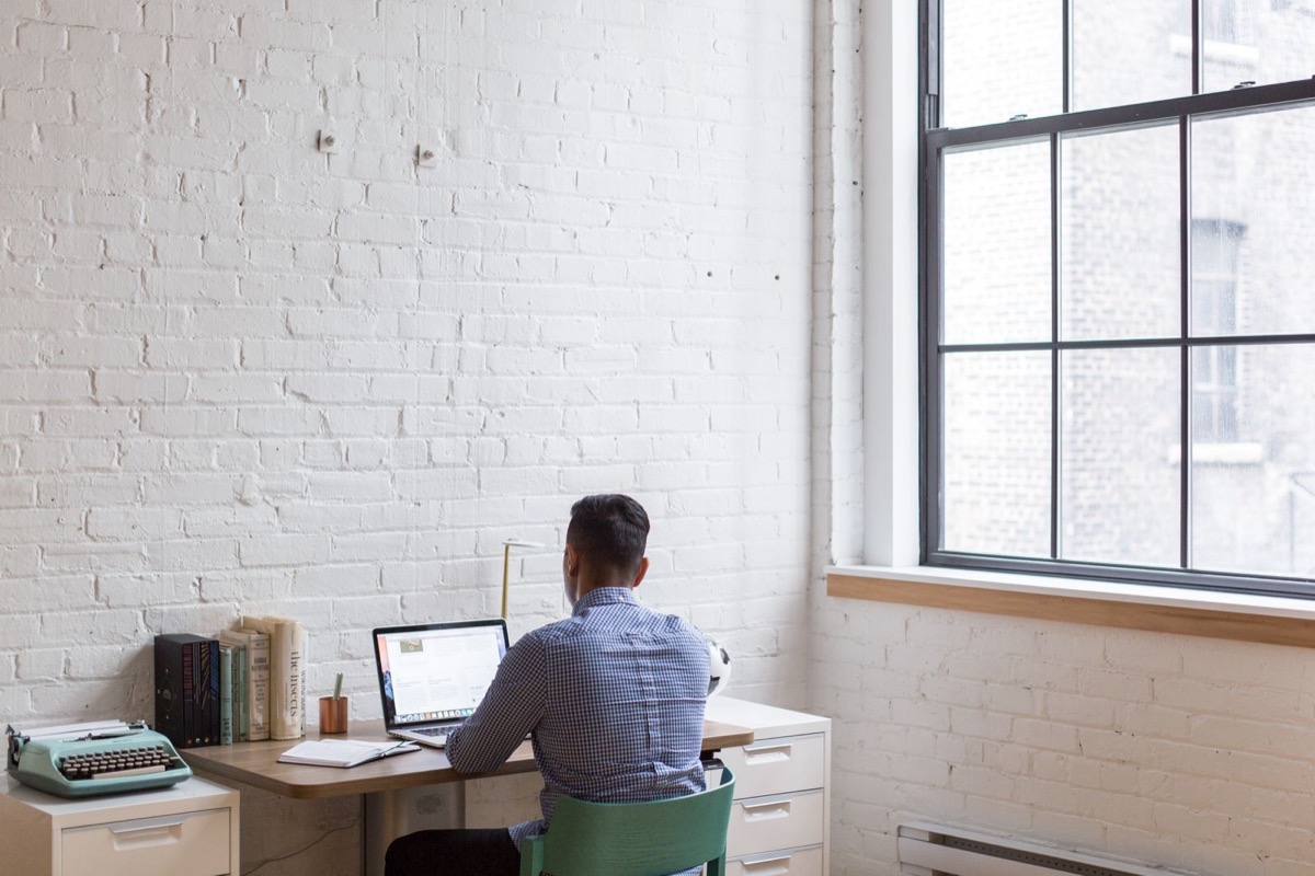 A person seen from behind working at a laptop at a desk in a bright room with a white brick wall