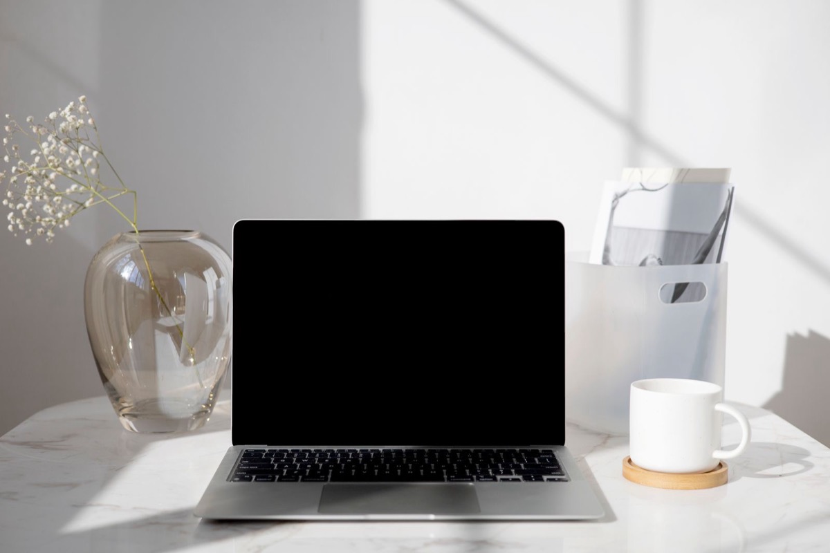 A laptop with a blank screen on a marble desk beside a glass vase and a white mug