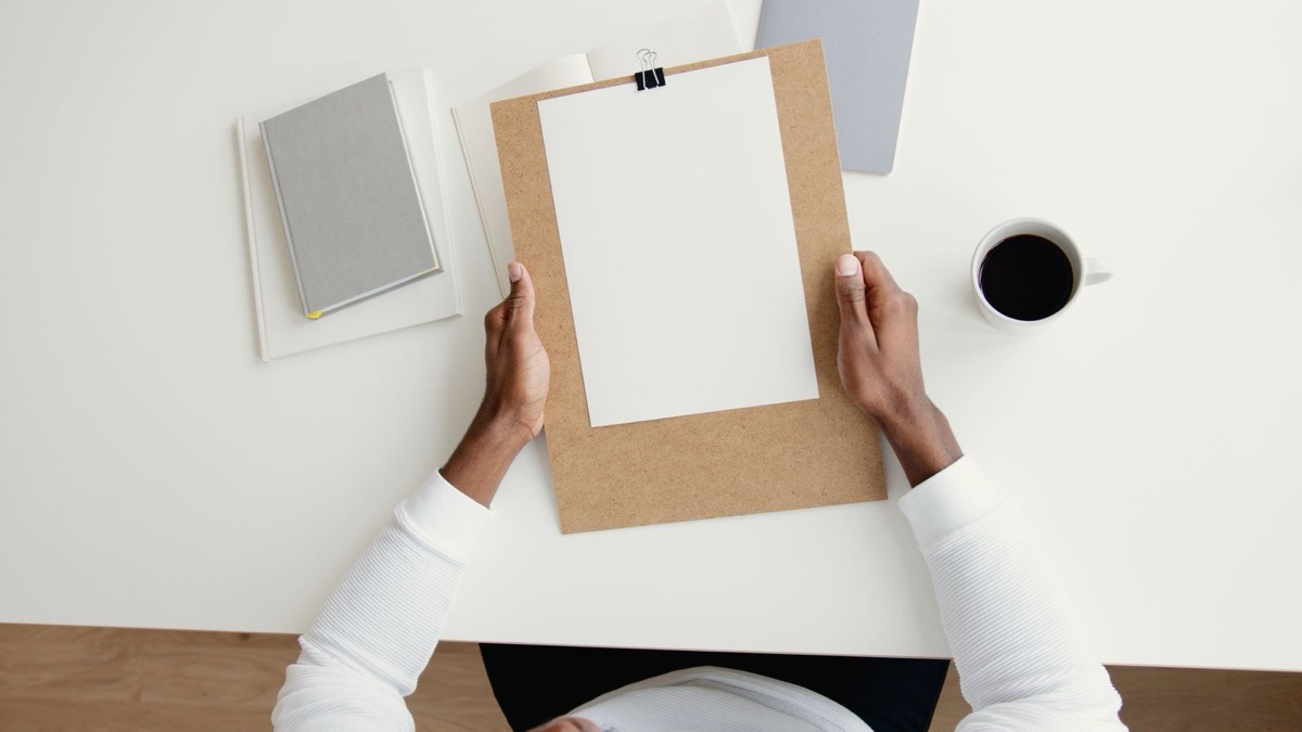 Hands holding a cardboard clipboard with a blank sheet at a white desk, with a grey notebook and a cup of coffee, seen from above