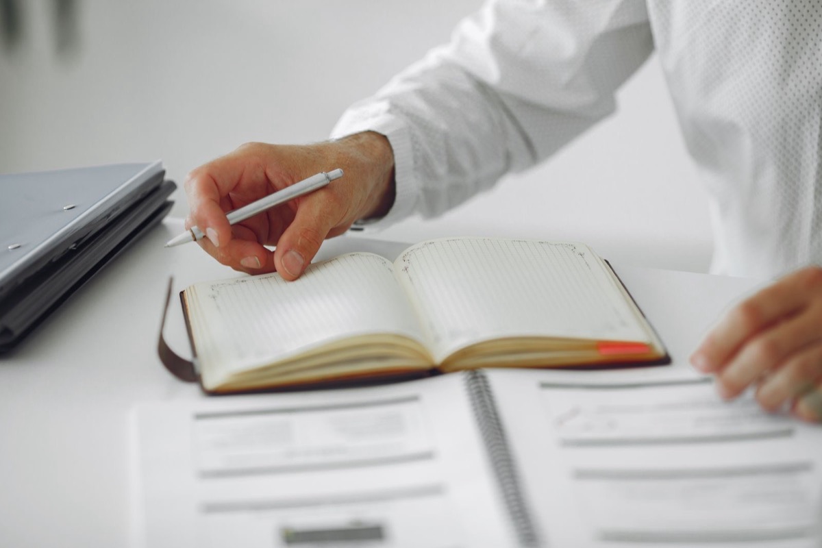 A hand holding a pen over an open diary, with printed forms below