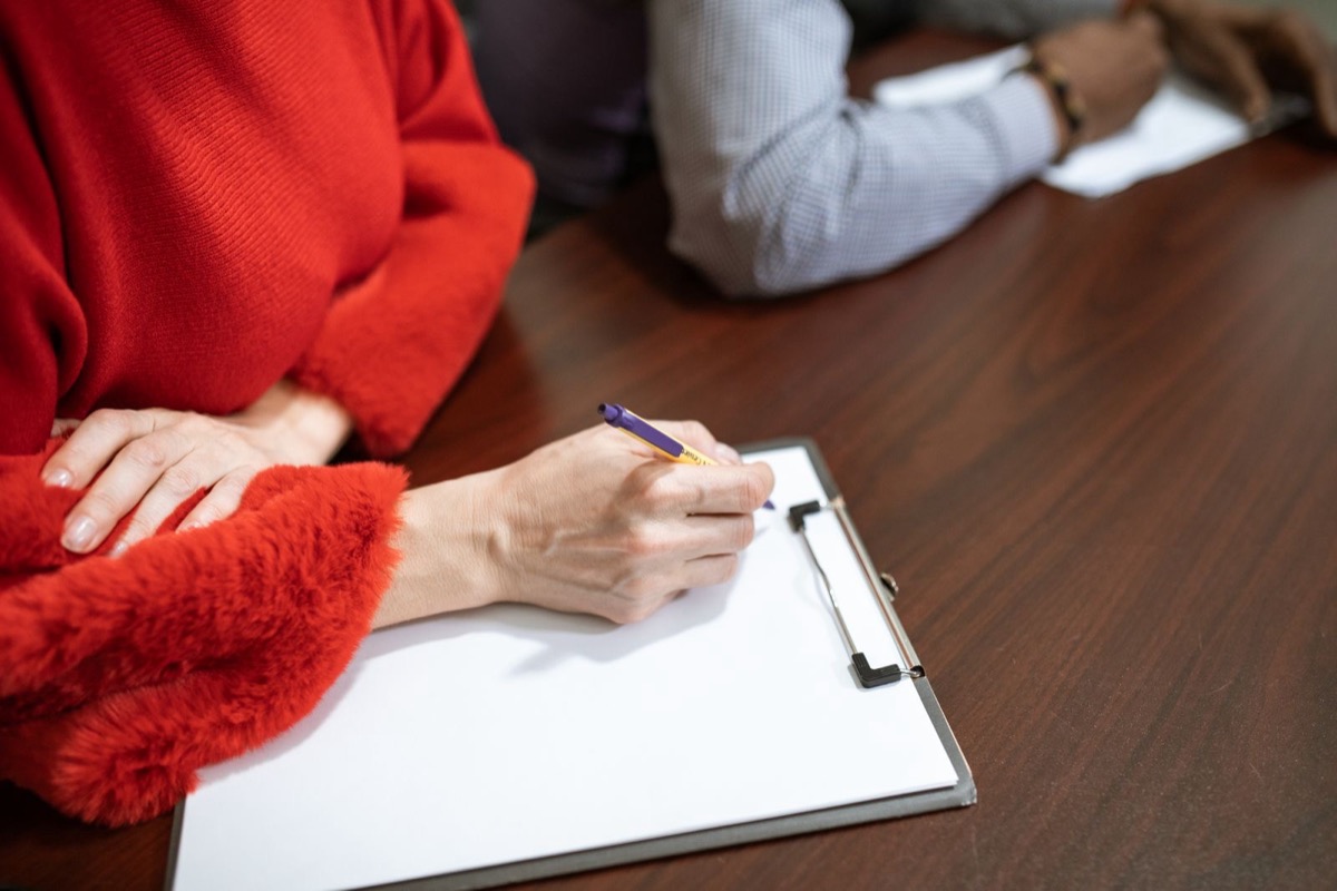 A hand holding a pen over a sheet on a clipboard at a dark wooden table