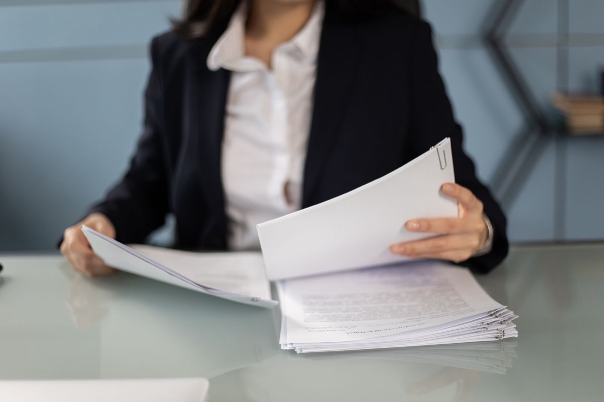 A woman in a dark jacket, seen from the chin down, holding printed pages above a stack of papers at a glass-topped desk