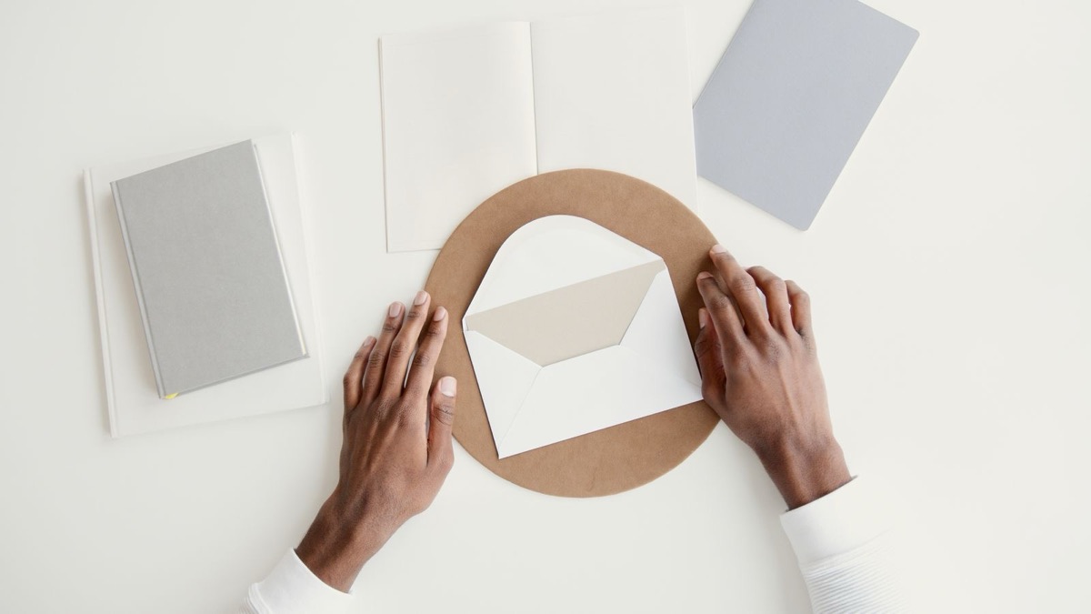 Two hands resting on a white envelope on a kraft disc, with a notebook and blank cards on a white desk