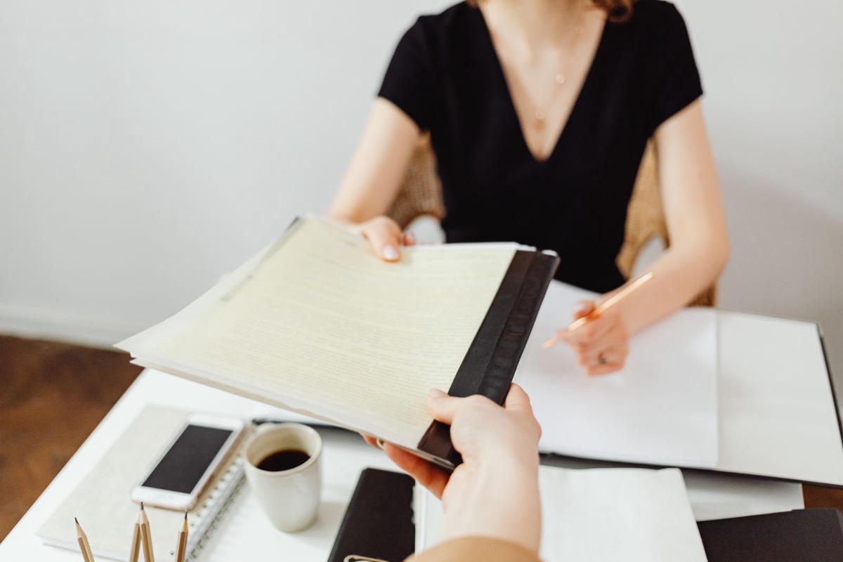 A hand passing a folder of typed papers across a desk to a seated person, with a phone, a cup and a notebook on the desk