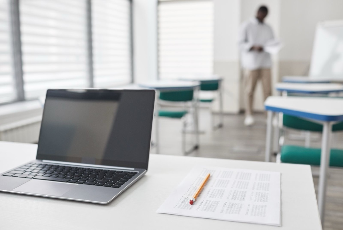 A laptop and a printed table with a pencil on a white desk in a bright room, a person walking in the background
