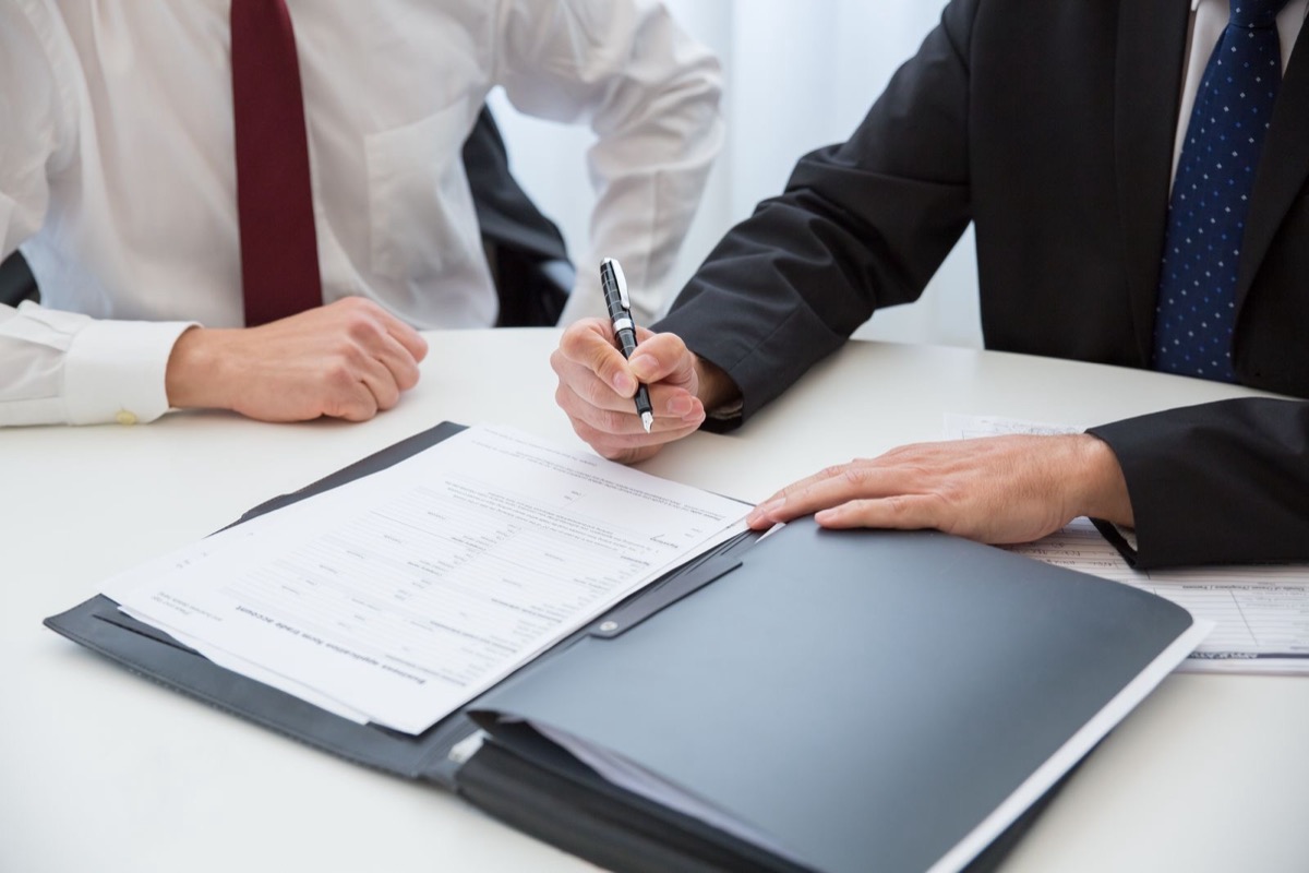Two people in business clothes at a white table, one signing papers in an open folder