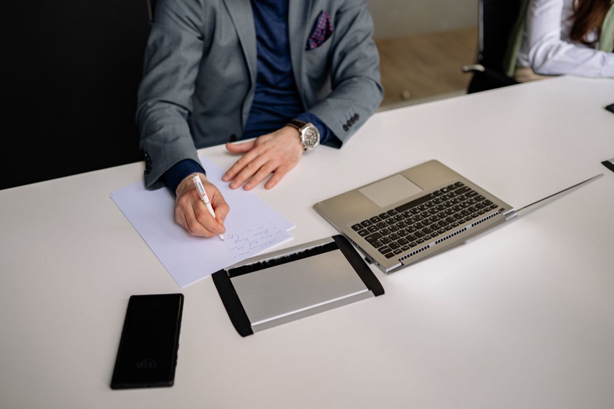 A person in a grey jacket writing on a sheet of paper at a white desk beside a laptop and a phone