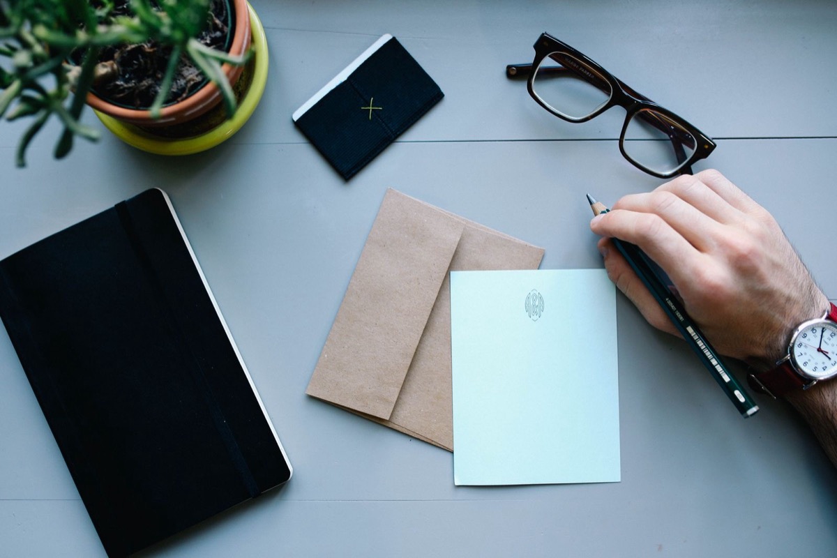 A kraft envelope and a pale card on a grey desk beside a closed notebook, glasses and a hand holding a pencil