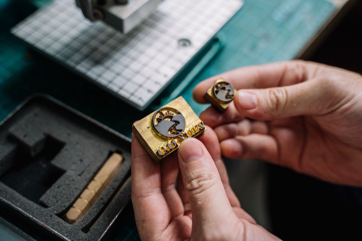 Two hands holding a small brass seal die above a foam-lined case
