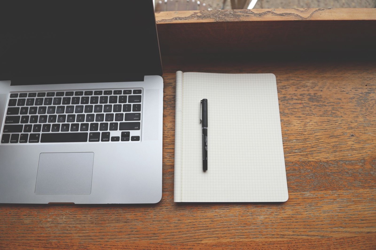 A laptop beside a squared notebook and a pen on a wooden desk