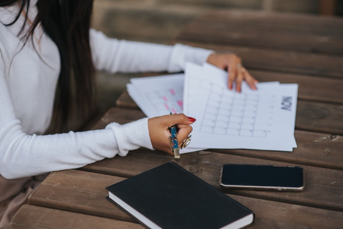 A hand holding a pen over printed calendar sheets on a wooden table, with a notebook and a phone