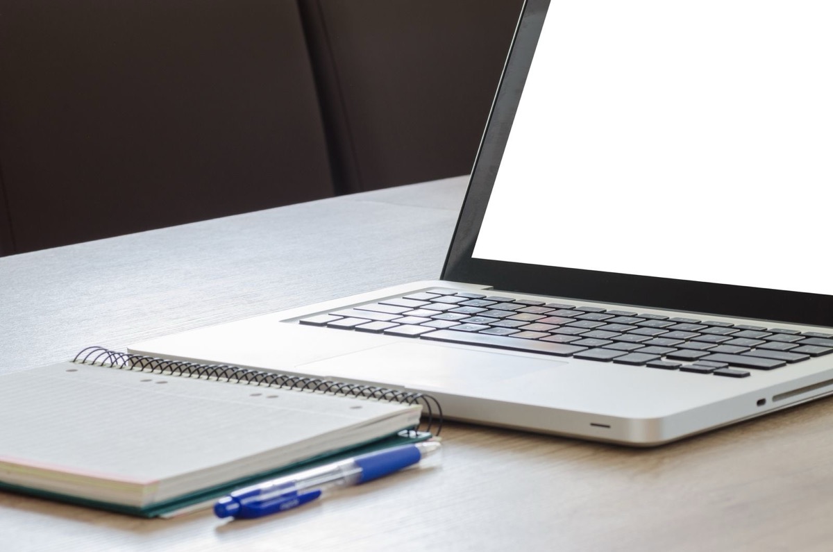 A laptop with a blank screen beside a spiral notebook and a pen on a desk